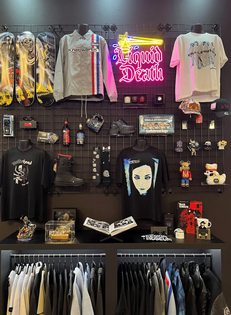 Global exhibition merchandise wall with artist apparel, Iron Maiden skateboard decks, accessories and collectibles beside a Liquid Death neon sign.